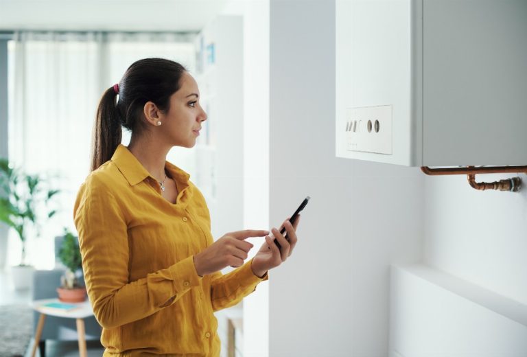 Femme réglant une chaudière gaz murale avec son smartphone dans un logement lumineux